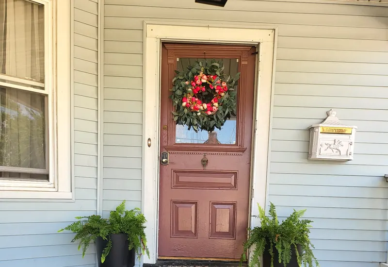 A red front door with a wreath on a porch with two potted ferns