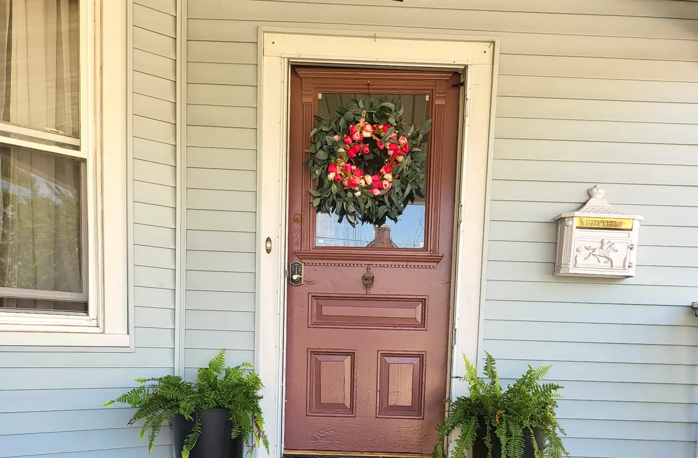 A red front door with a wreath on a porch with two potted ferns