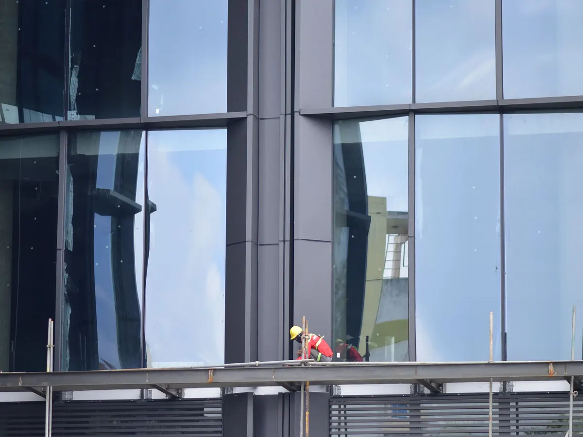 A worker in a hard hat on a ledge below a tall glass curtain wall