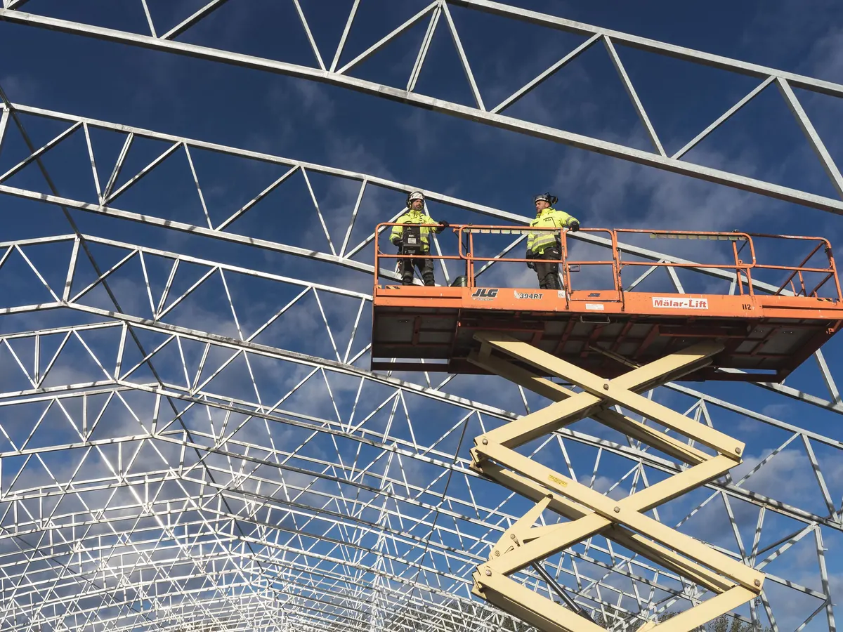Two workers in safety vests on a scissor lift under a steel roof frame