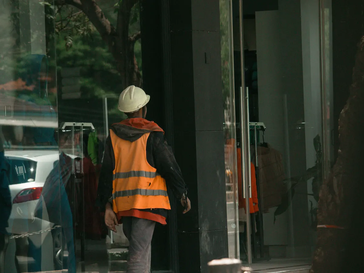 A worker in a hard hat and safety vest at a glass storefront entrance