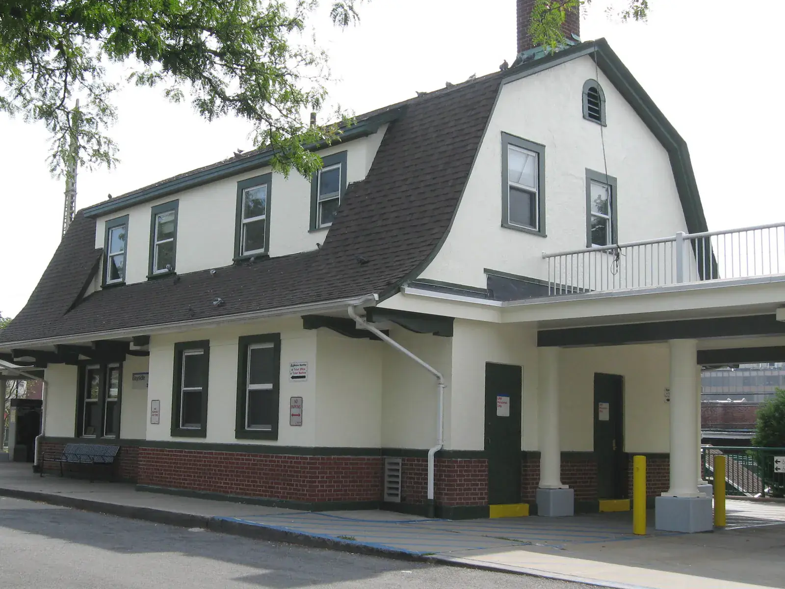The white Bayside train station house with a dark gambrel roof and green trimmed windows under a pale sky