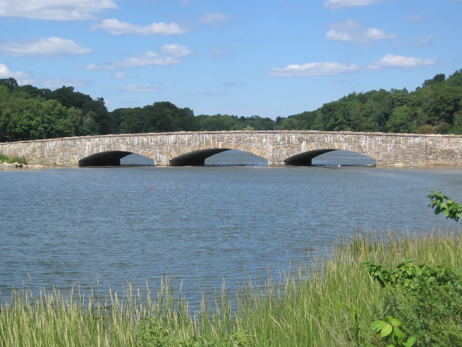 A three-arch stone bridge over blue water in Darien with marsh grass in the foreground and wooded hills under a blue sky