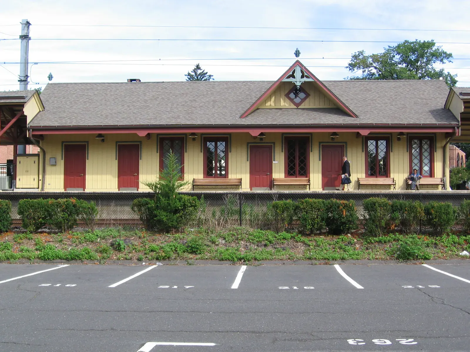 The New Canaan train station, a yellow wooden building with red doors and a grey roof, seen across a hedge and a parking area