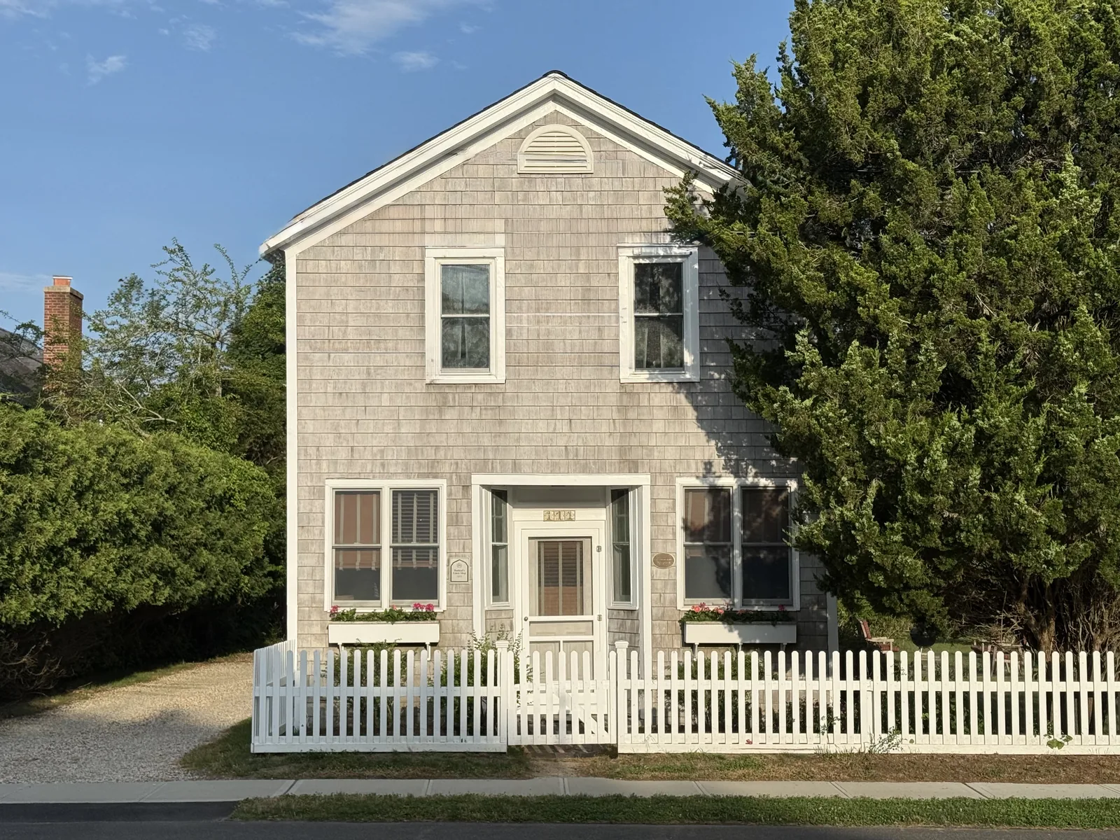 A grey cedar-shingled house with white windows, window boxes and a white picket fence in Quogue under a blue sky