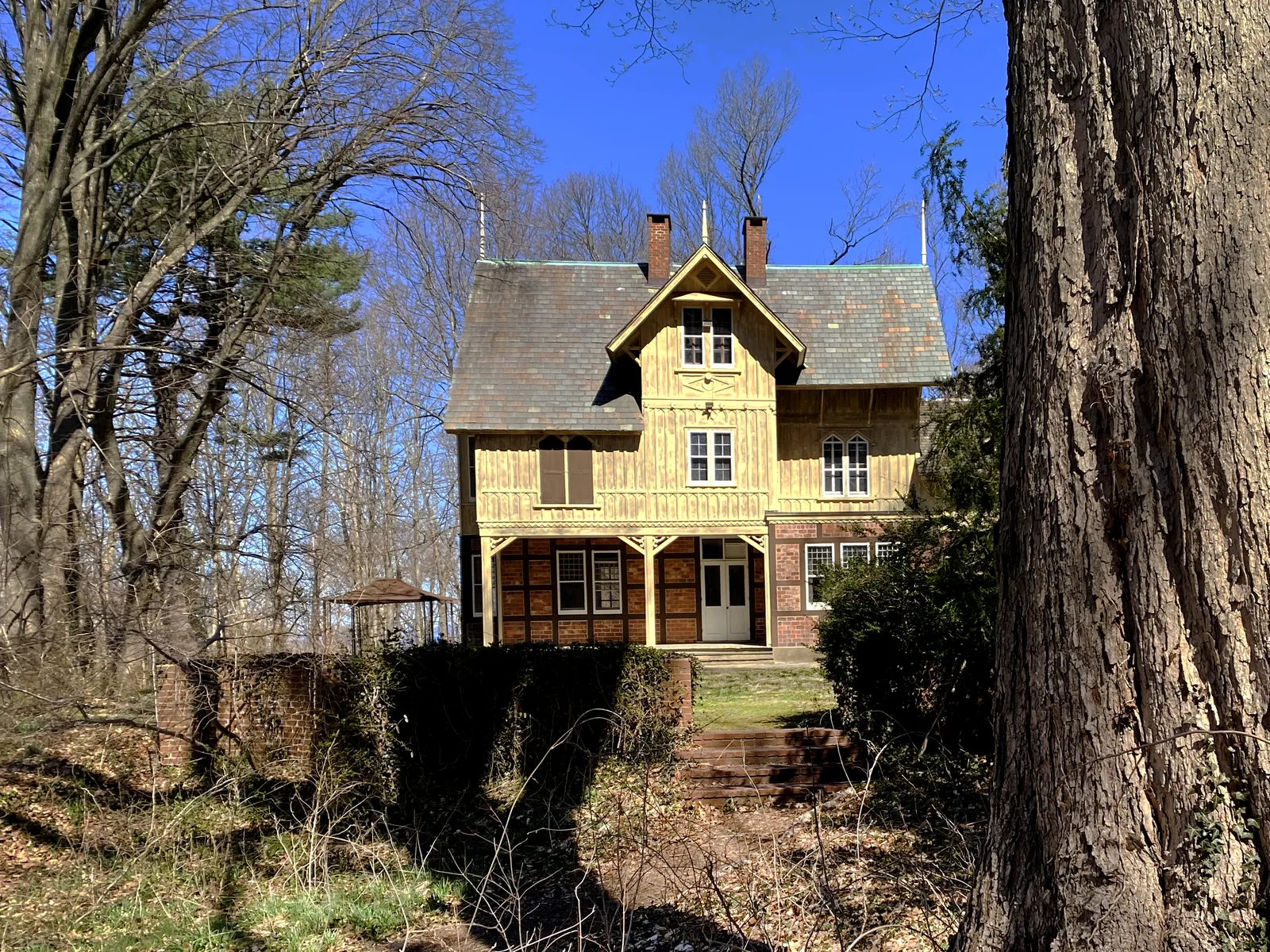 Yellow 1862 cottage with a steep gabled roof among tall trees on the Nassau County Museum of Art grounds in Roslyn Harbor, NY