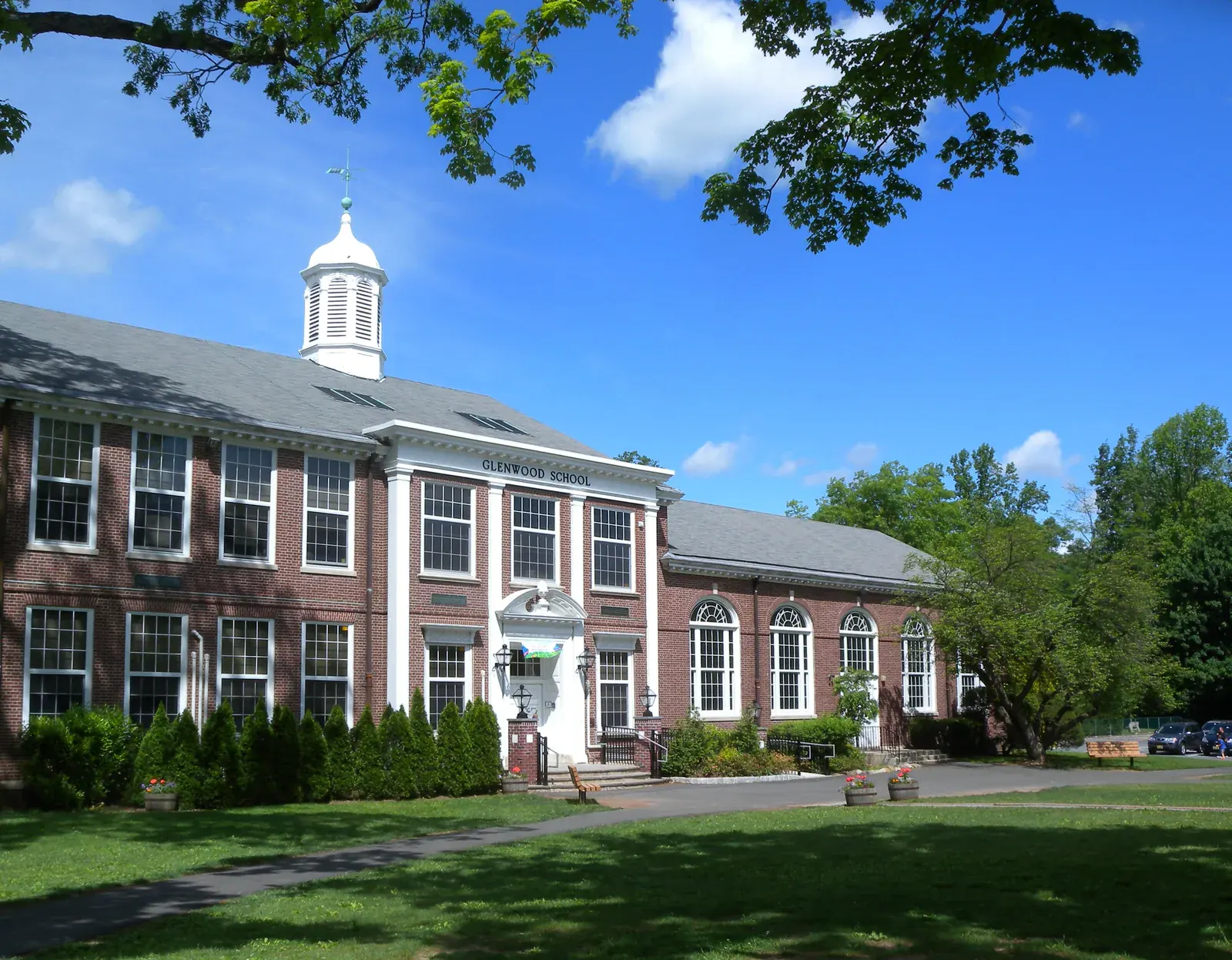 A red-brick school with white trim and a white cupola behind a lawn and trees in Short Hills under a bright blue sky