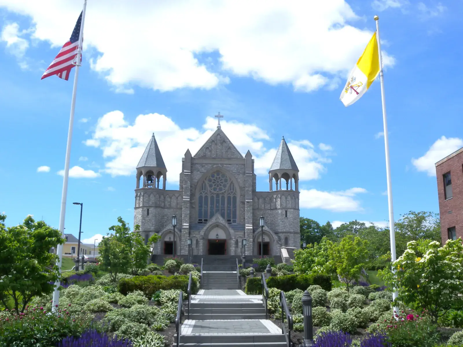 A grey stone church with twin towers at the top of garden steps in Summit, between two flagpoles under a blue sky with white clouds