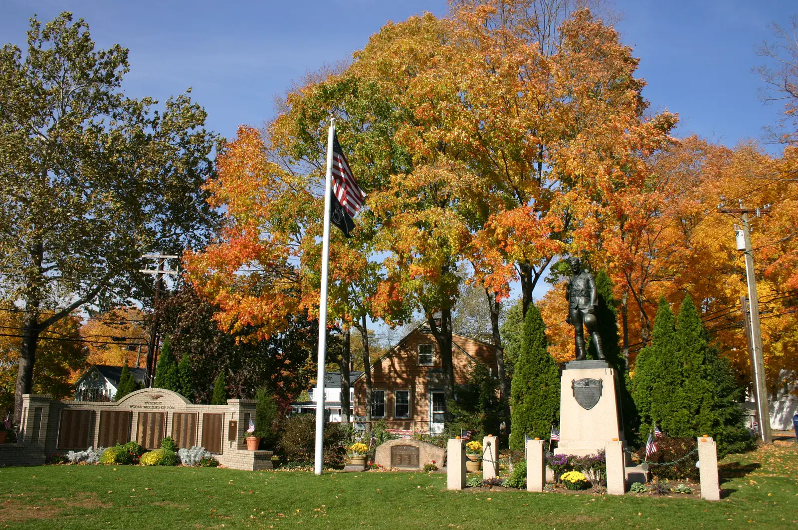 A town green in Westport with a flagpole, a soldier statue and memorial walls under orange autumn trees and a deep blue sky
